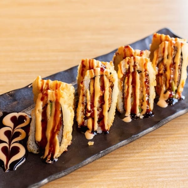 Five pieces of deep-fried sushi rolls drizzled with two sauces on a black rectangular plate.