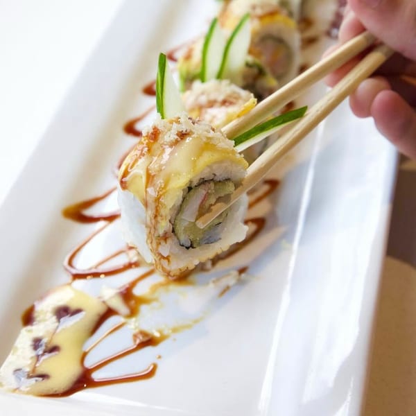 Chopsticks picking up a sushi roll with sauce drizzle and cucumber garnish on a white plate.