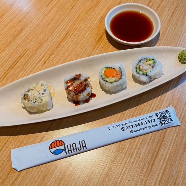 Four sushi rolls on a white plate with soy sauce and a Kaja branded chopstick wrapper.