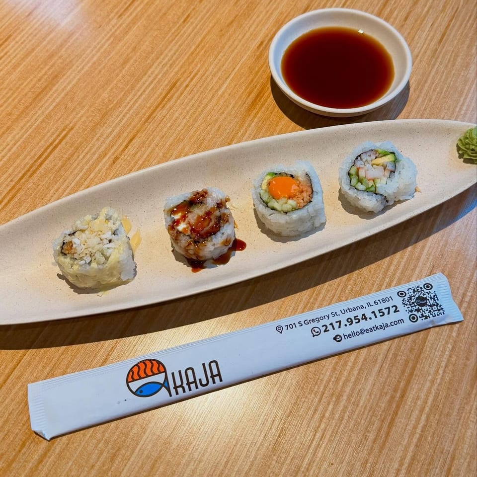 Four sushi rolls on a white plate with soy sauce and a Kaja branded chopstick wrapper.