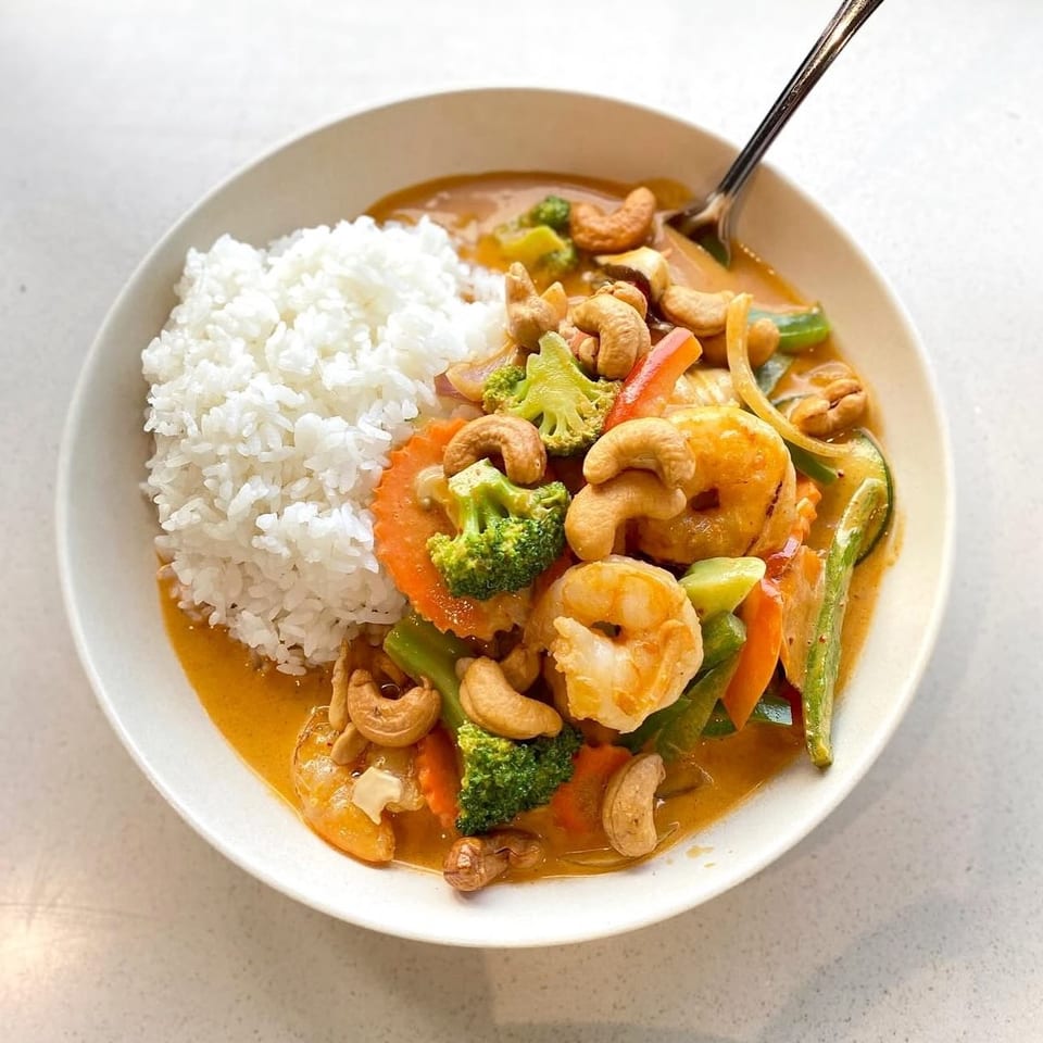 Shrimp curry with broccoli, carrots, cashews, and rice in a white bowl.