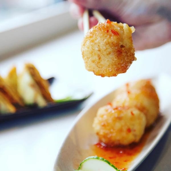 Golden fried appetizer ball held by chopsticks, with more on a plate with sauce and cucumber.