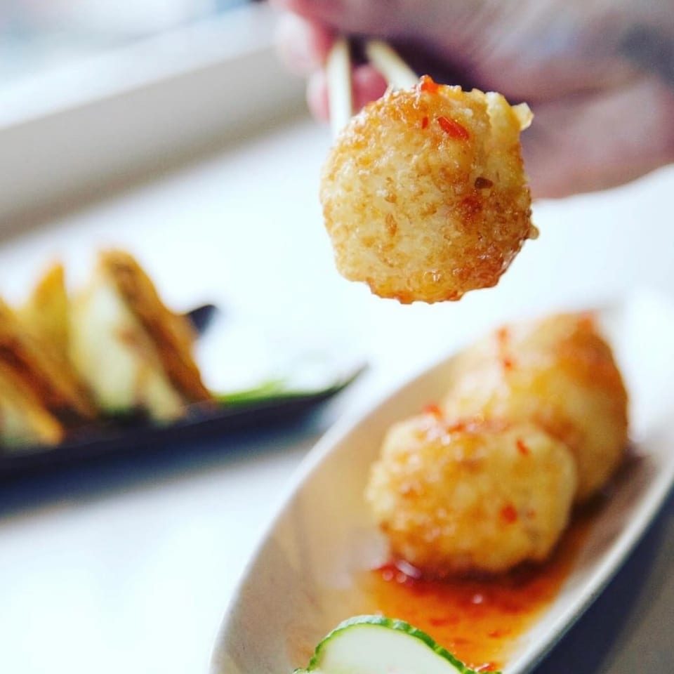 Golden fried appetizer ball held by chopsticks, with more on a plate with sauce and cucumber.