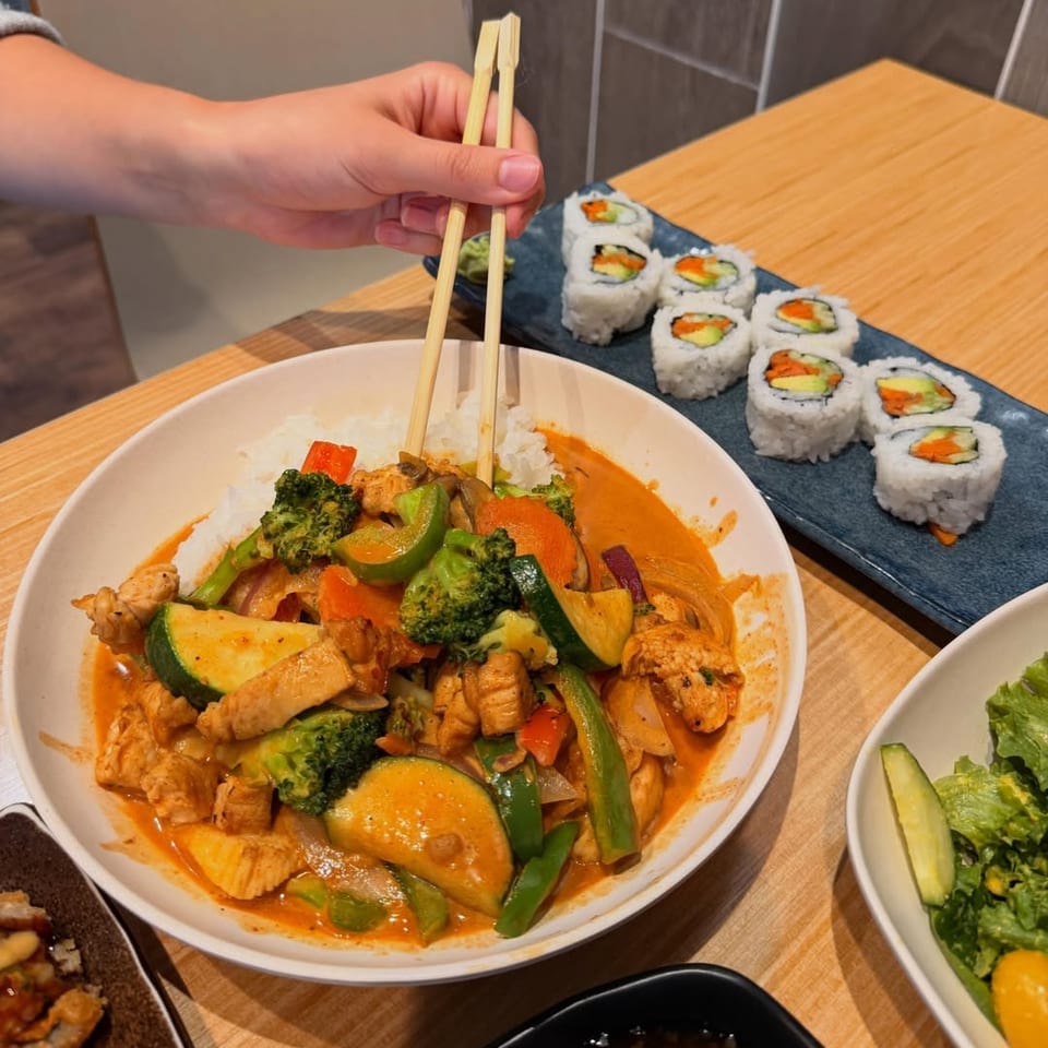 A hand holding chopsticks over a bowl of red curry with chicken and vegetables, with sushi rolls nearby.