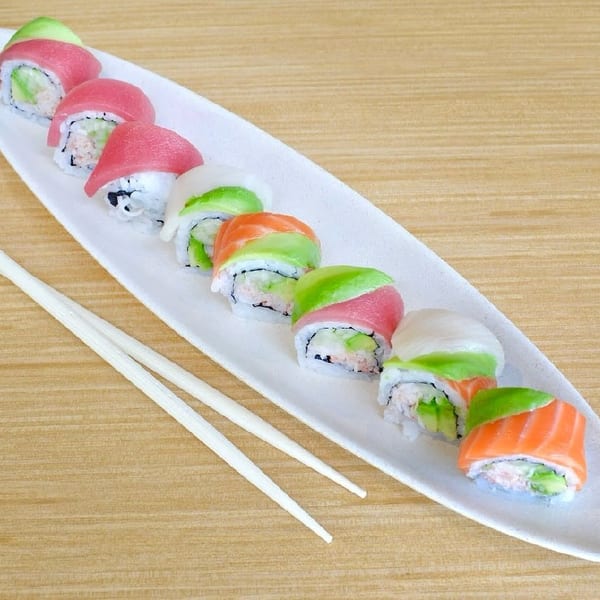 A long white plate holds a row of colorful sushi rolls with chopsticks on a wooden table.
